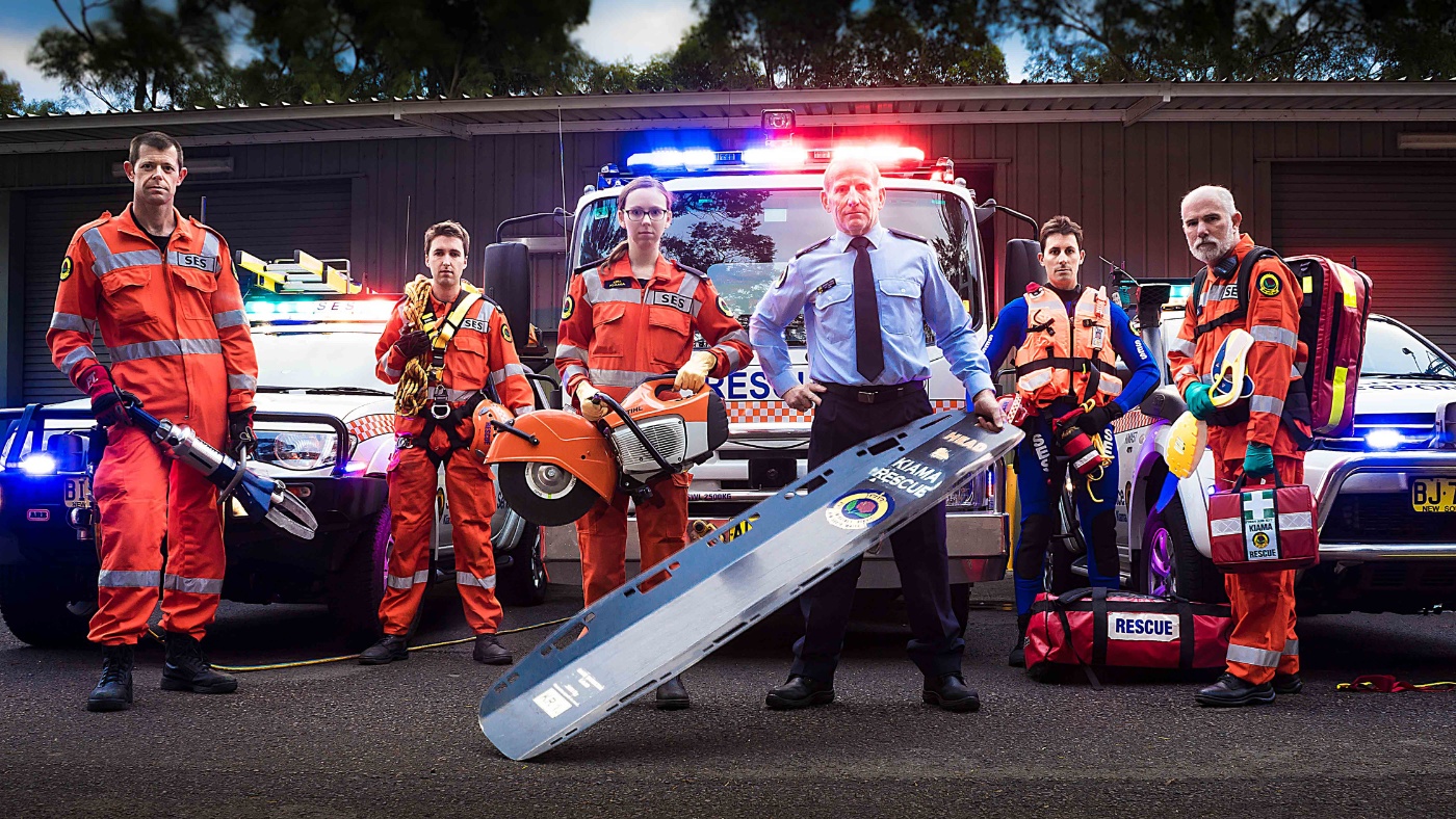 Six NSW SES members in orange uniforms holding rescue tools in front of a NSW SES truck and vehicles..