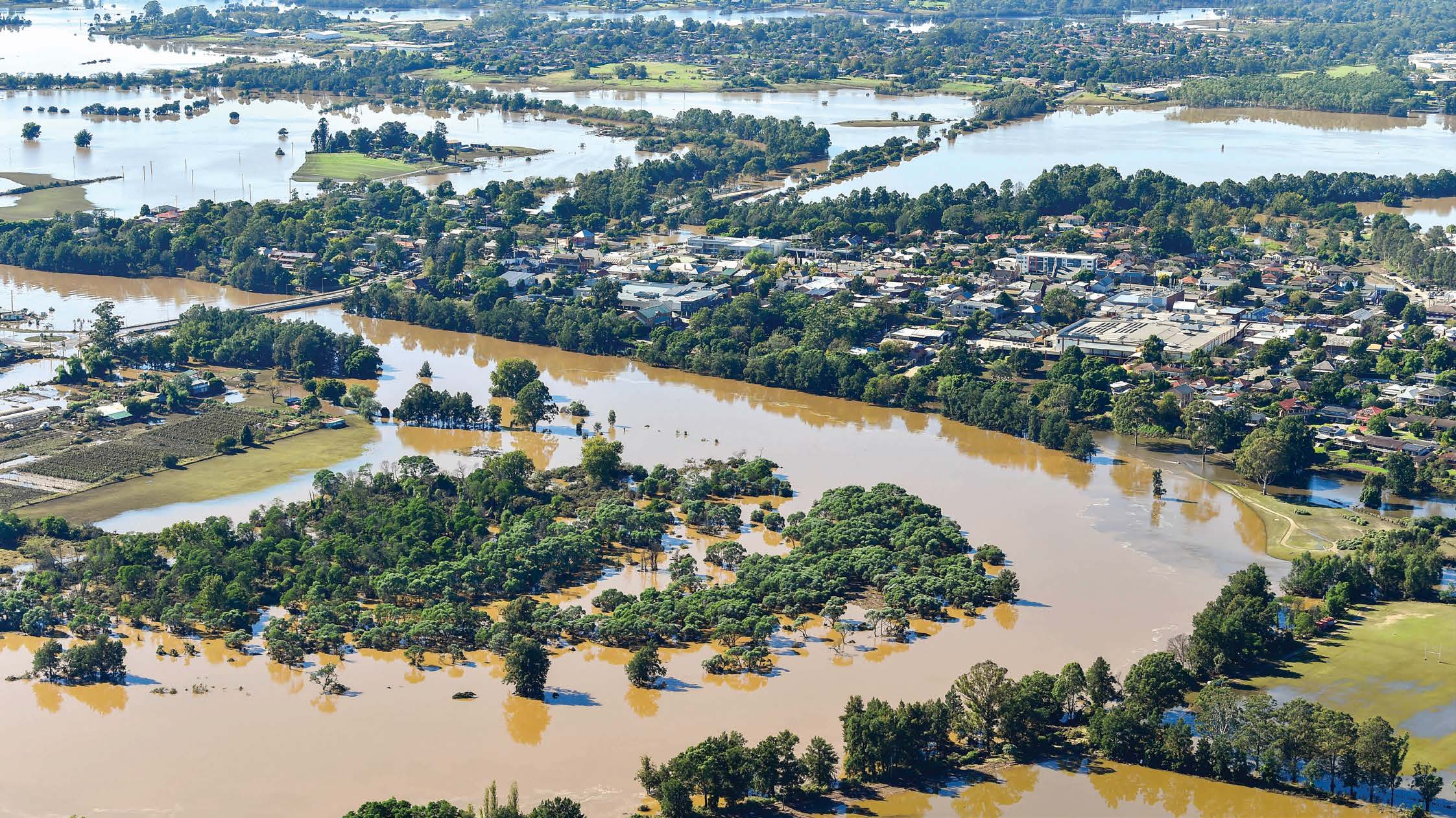 Flooded landscape in the Hawkesbury-Nepean area of NSW, with fields and trees partially submerged under murky floodwater.